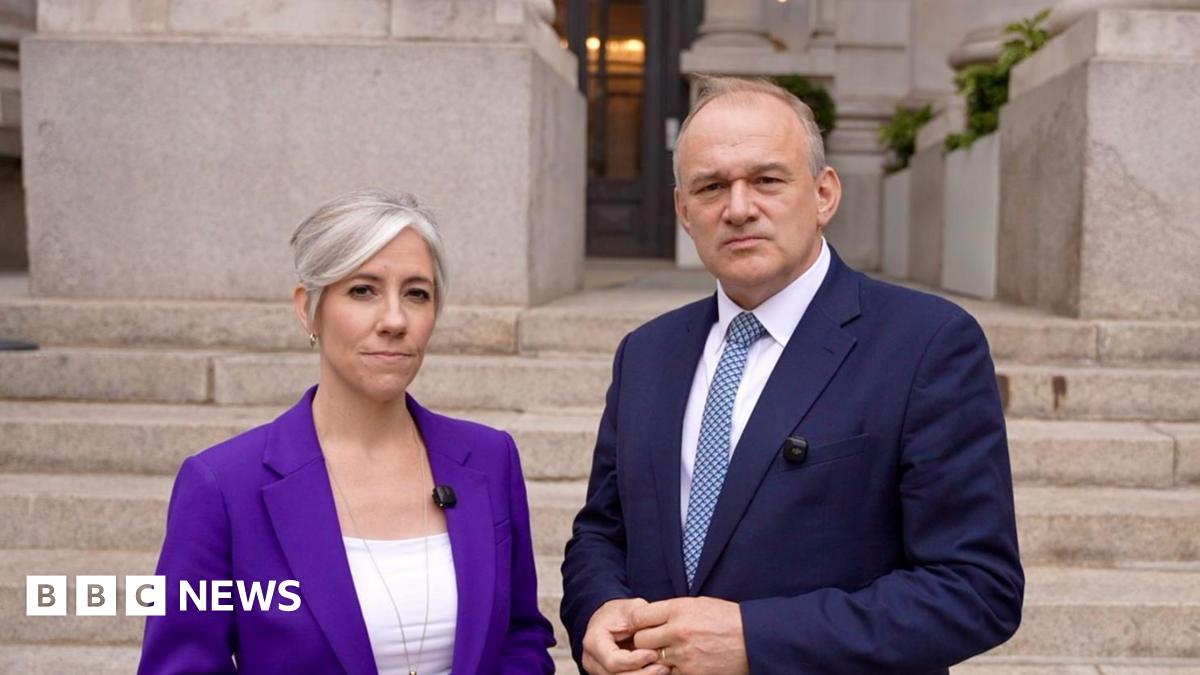 Sir Ed Davey and Daisy Cooper stand outside the Bank of England. Ed Davey wears a dark blue suit, white shirt, and blue patterned tie; Daisy Cooper wears a purple blazer over a white top. Behind them are stone steps and the grand entrance of the Bank of England