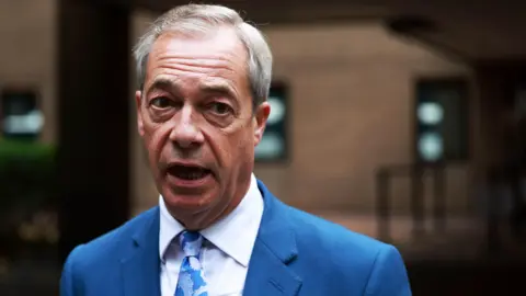 EPA Nigel Farage is pictured while speaking outdoors in front of a blurred brick building with windows. He wears a blue suit jacket, white dress shirt, and blue patterned tie.