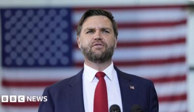 A man in a suit. He is wearing a red tie and has the US flag pinned on his lapel. Behind him is the US flag.