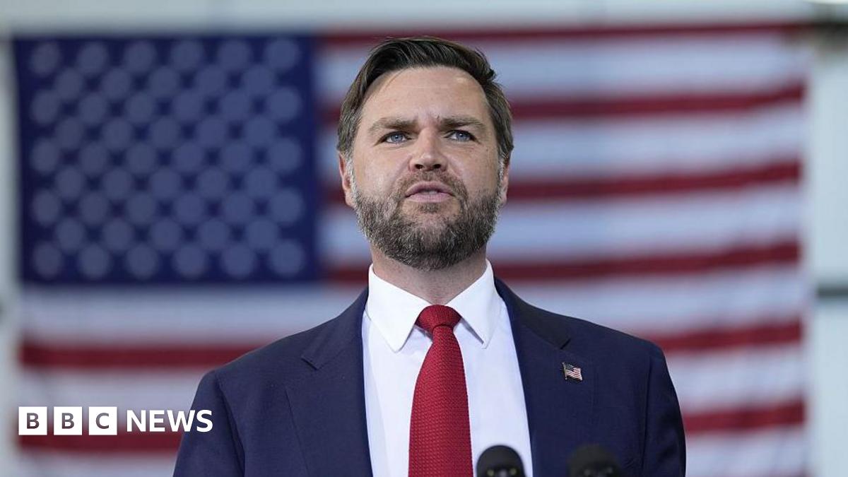 A man in a suit. He is wearing a red tie and has the US flag pinned on his lapel. Behind him is the US flag.