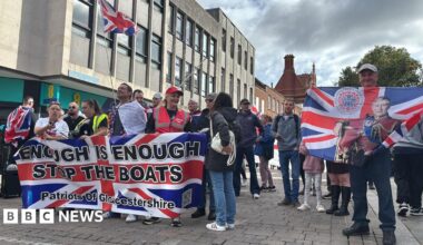 A crowd of people holding Union flags on a pedestrianised street. The largest flag is printed with the words "Enough is Enough, Stop the boats, Patriots of Gloucestershire". One man holds a Union flag with King Charles II printed on it.