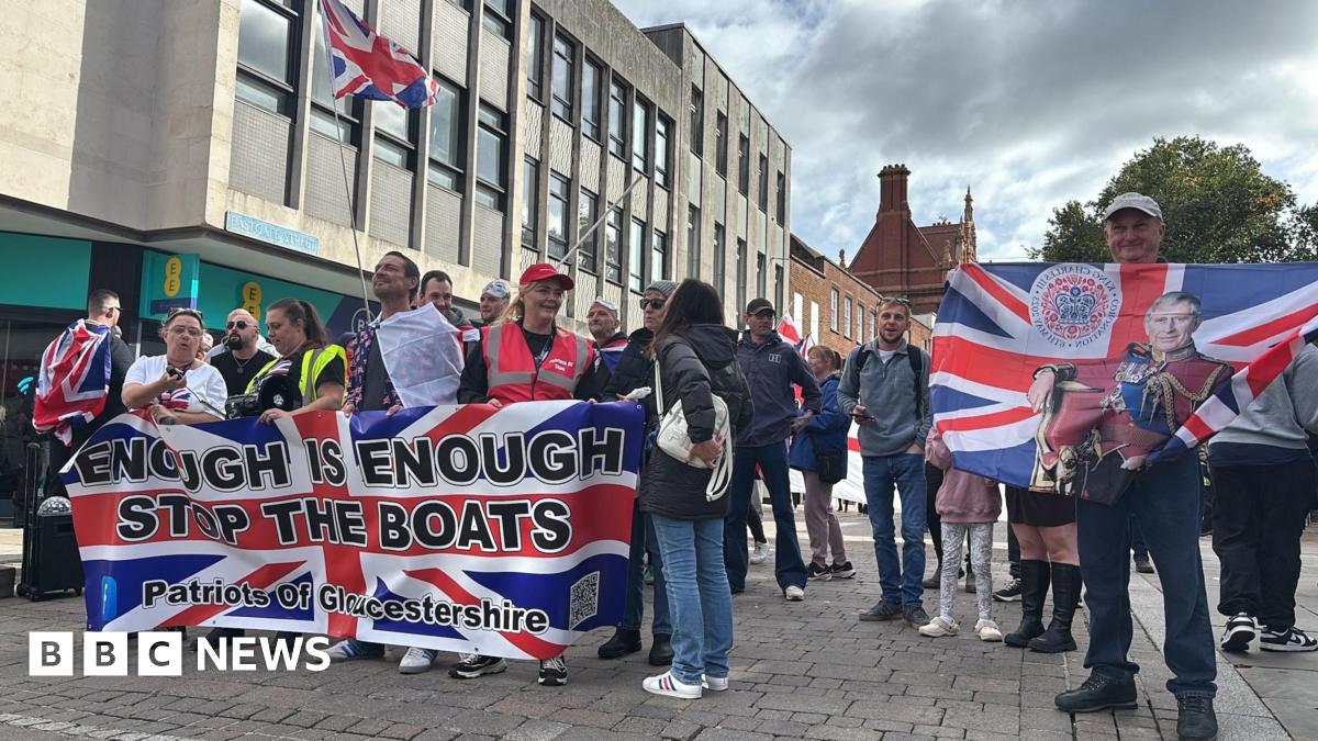 A crowd of people holding Union flags on a pedestrianised street. The largest flag is printed with the words "Enough is Enough, Stop the boats, Patriots of Gloucestershire". One man holds a Union flag with King Charles II printed on it.