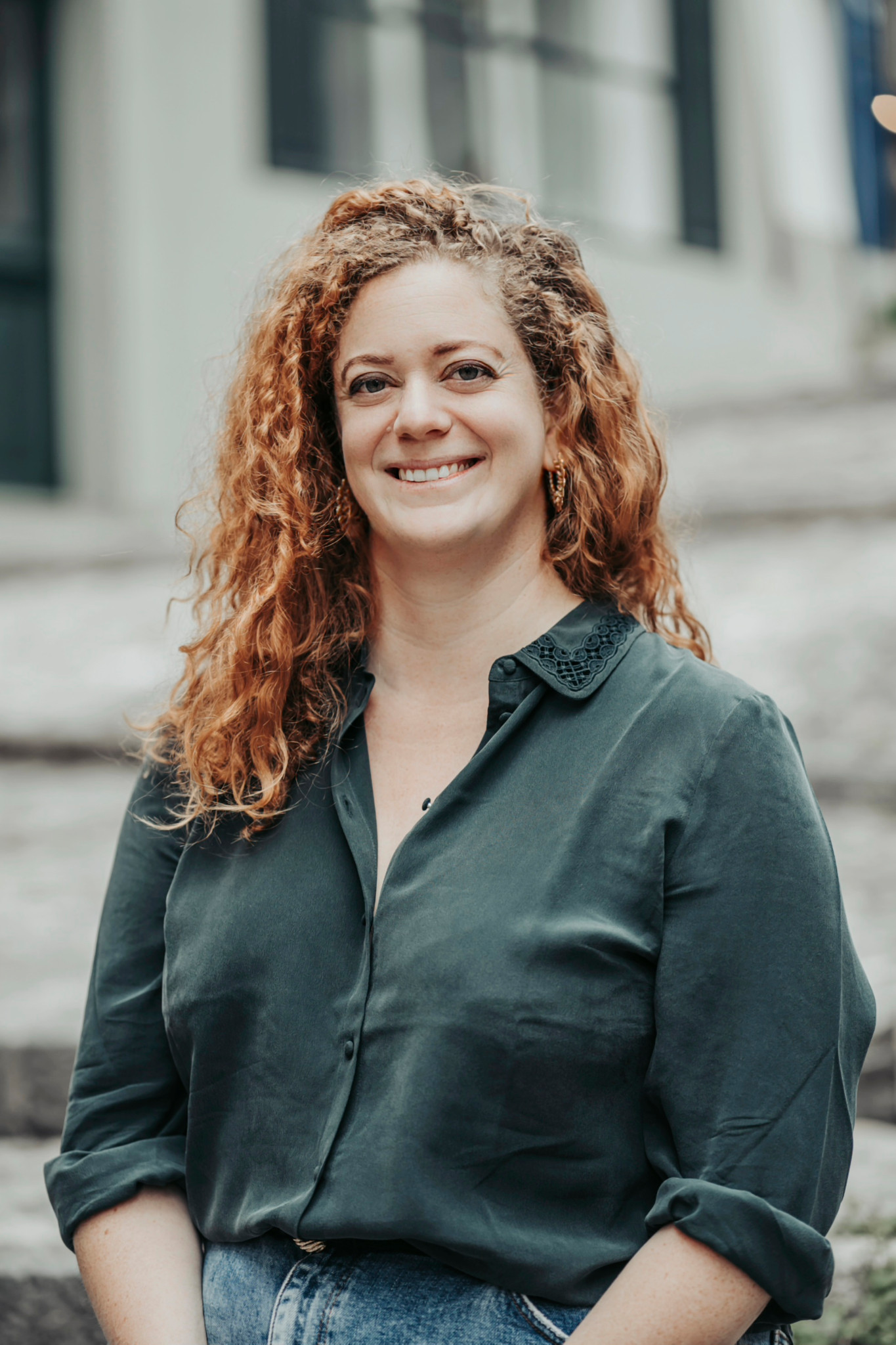 Frau mit lockigen, roten Haaren lächelt in die Kamera, trägt ein grünes Hemd, steht vor einer verschwommenen Treppenkulisse.