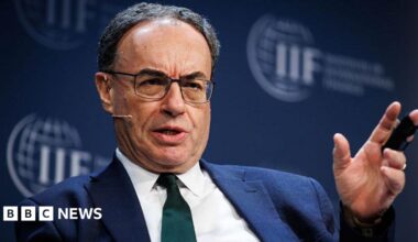Andrew Bailey speaking on stage while sitting down at an International Institute of Finance event, the logo of which is visible on a backdrop behind Bailey. The governor has a serious expression, is mid-saying something, has a head mic, and is wearing a dark green tie with a blue jacket and white shirt. He is gesturing with his left hand as he speaks.