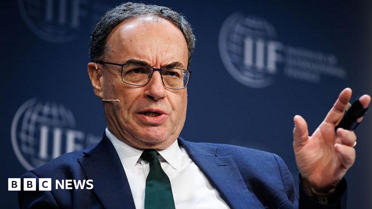 Andrew Bailey speaking on stage while sitting down at an International Institute of Finance event, the logo of which is visible on a backdrop behind Bailey. The governor has a serious expression, is mid-saying something, has a head mic, and is wearing a dark green tie with a blue jacket and white shirt. He is gesturing with his left hand as he speaks.
