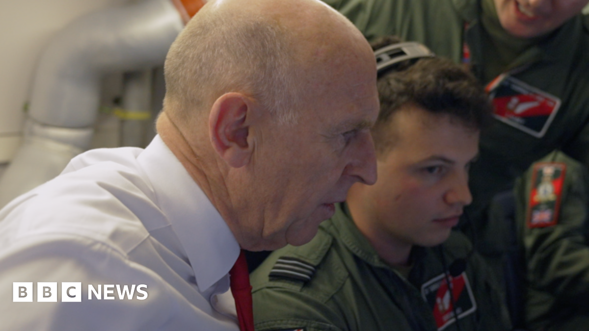 Defence Secretary John Healey in side profile, wearing a white shirt and red tie, alongside RAF crew in uniform