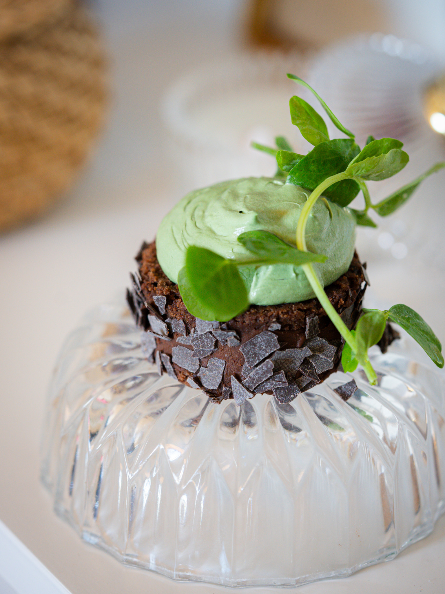 Dessert chocolaté surmonté de crème verte et décoré de feuilles, présenté sur une base en verre texturé. Dessert chocolaté surmonté de crème verte et décoré de feuilles, présenté sur une base en verre texturé.