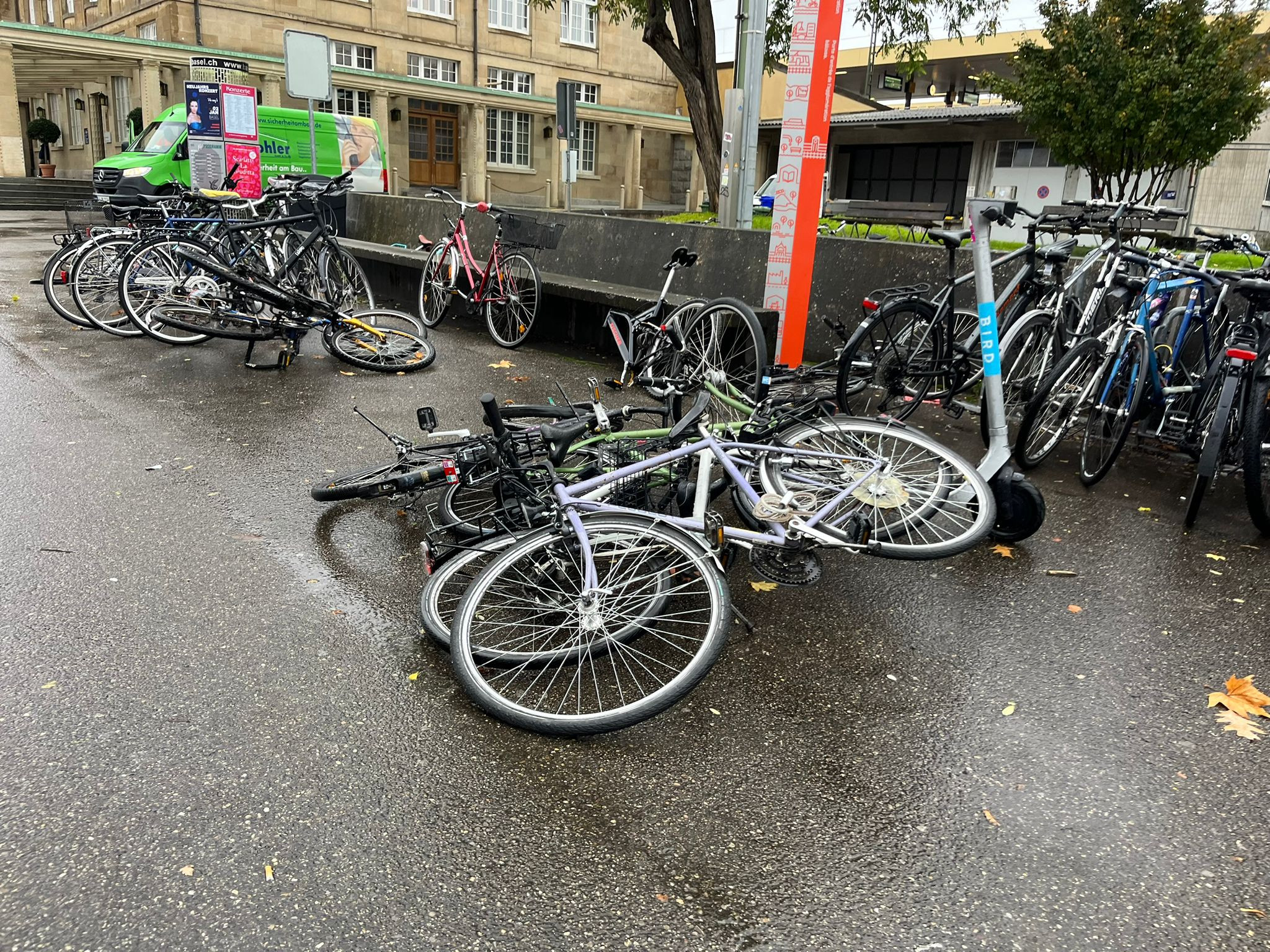 Umgekippte Fahrräder auf nasser Strasse in der Nähe eines Gebäudes, mit parkenden Autos im Hintergrund und einem elektronischen Informationsschild.