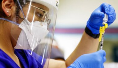 A woman wearing glasses, a face mask and a visor with blue surgical gloves holds a syringe and a small bottle.