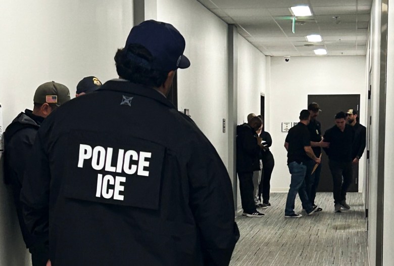 Immigration court hallway. An ICE officer stands as others detain an immigrant at the end of the hall.