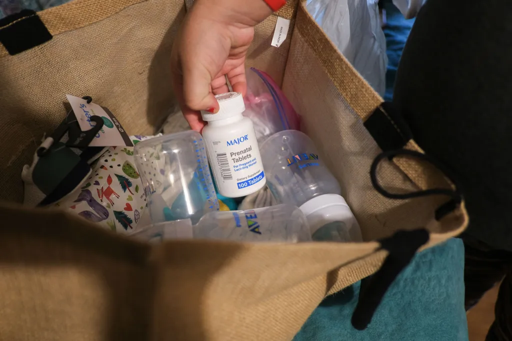 A close-up of a hand holding a white bottle labeled “Prenatal Tablets” over a bag filled with baby bottles and other supplies.