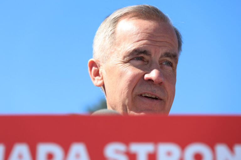 A close-up of Mark Carney's face against a backdrop of blue sky. A portion of a red-and-white lectern sign is visible in the lower foreground.