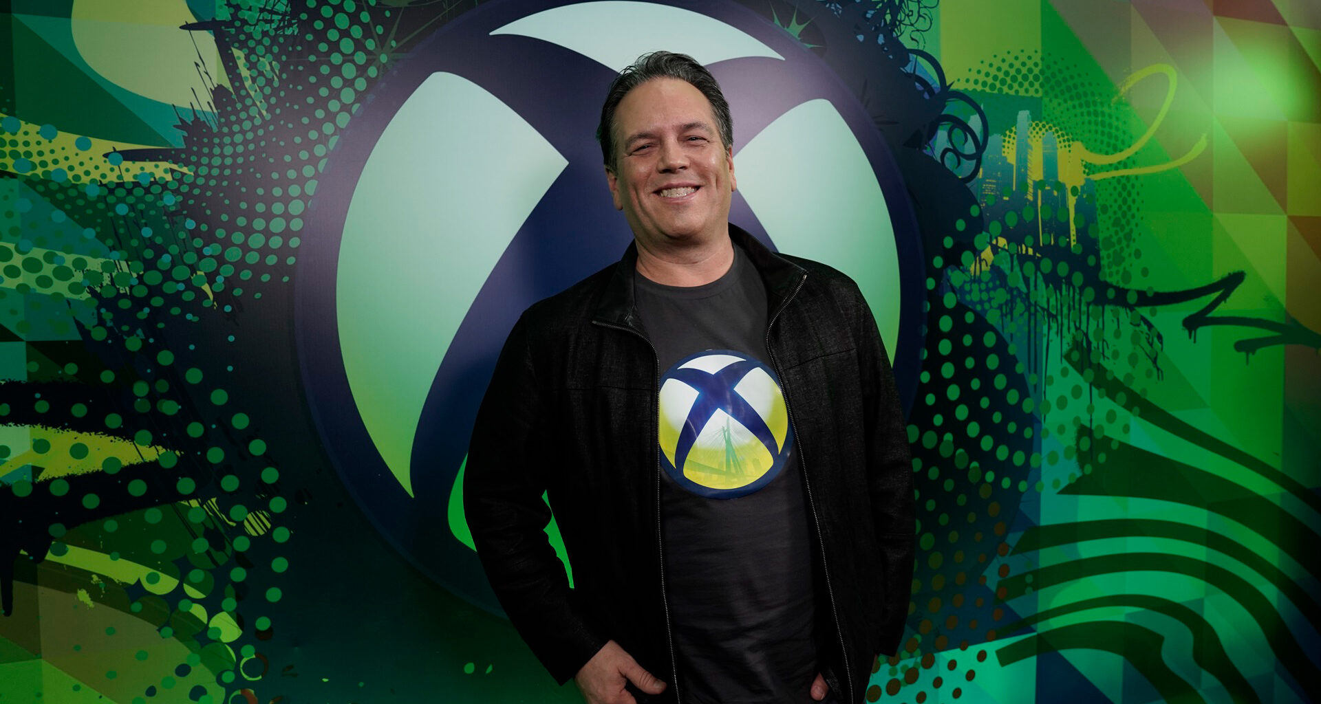 Phil Spencer standing in front of Xbox logo wall wearing a t-shirt with the Xbox symbol.