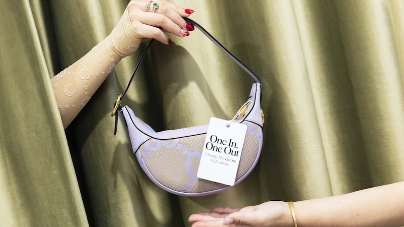 A female hand lifts a purple handbag from behind a curtain and hands if off to an outstretched arm. The handbag bears the label "One in, one out. Change the future of fashion."