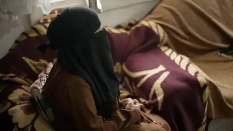 Profile of a woman sitting on a sofa. She is wearing a brown, long-sleeved dress with thin white stripes and a black headscarf which covers her face. Only part of one eye can be seen.