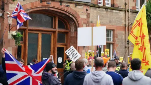 PA Media Groups of protesters outside a hotel housing asylum seekers in Perth. A Union Jack flag is being raised in the foreground as is a Lion Rampant. In the background, a protesters is holding a placard with the phrase 'no hate, no fear, refugees are welcome here'.