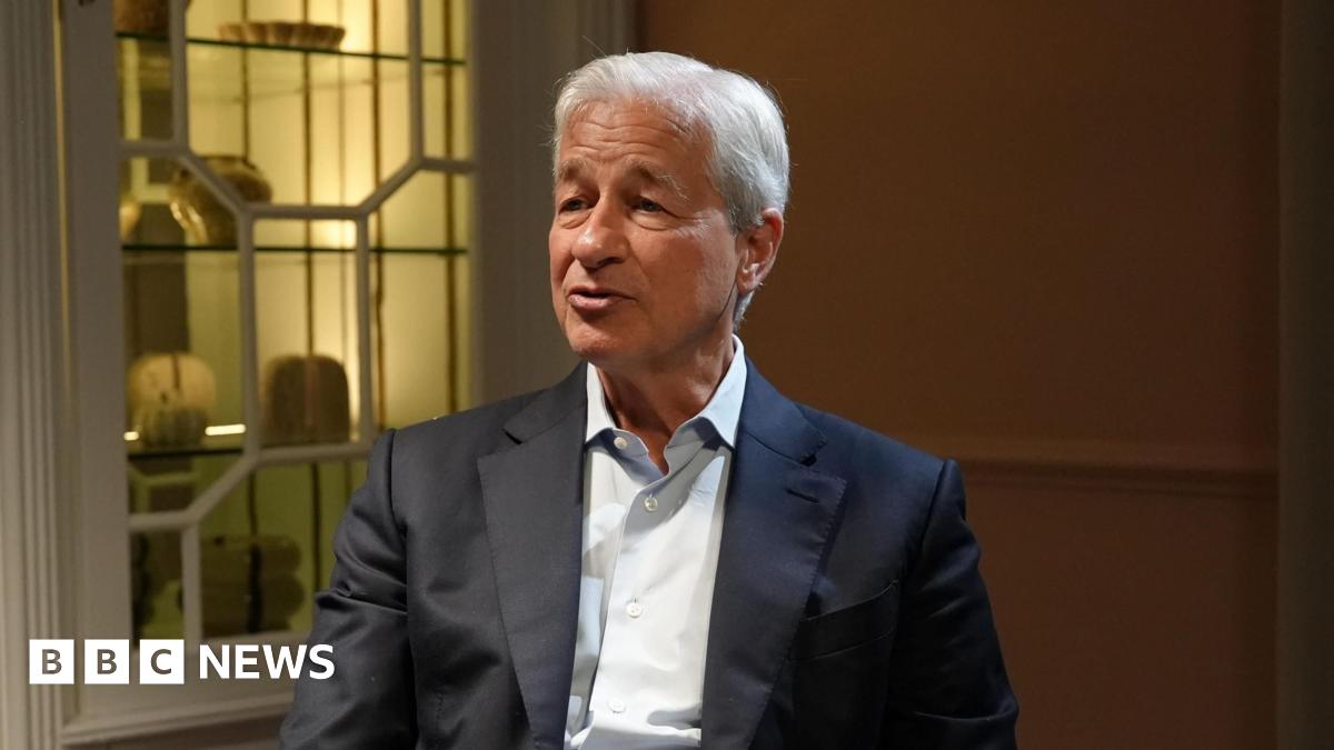 BBC Business editor Simon Jack sits opposite Jamie Dimon as he interviews him near a fire place surrounded by TV cameras