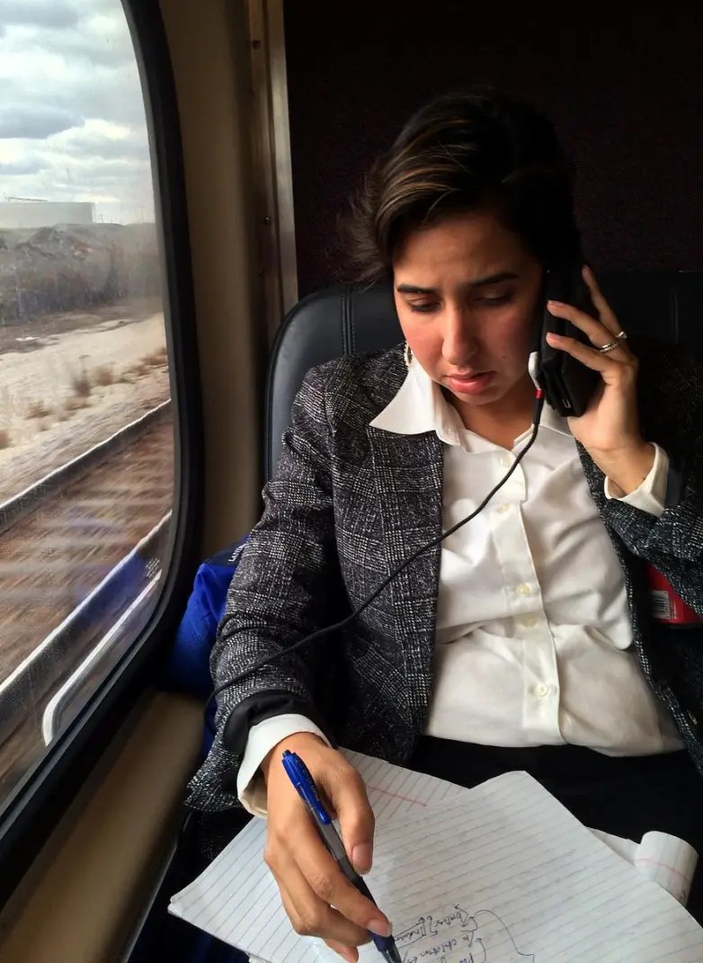 A woman wearing a suit speaks on the phone and takes notes, seated next to a train window.