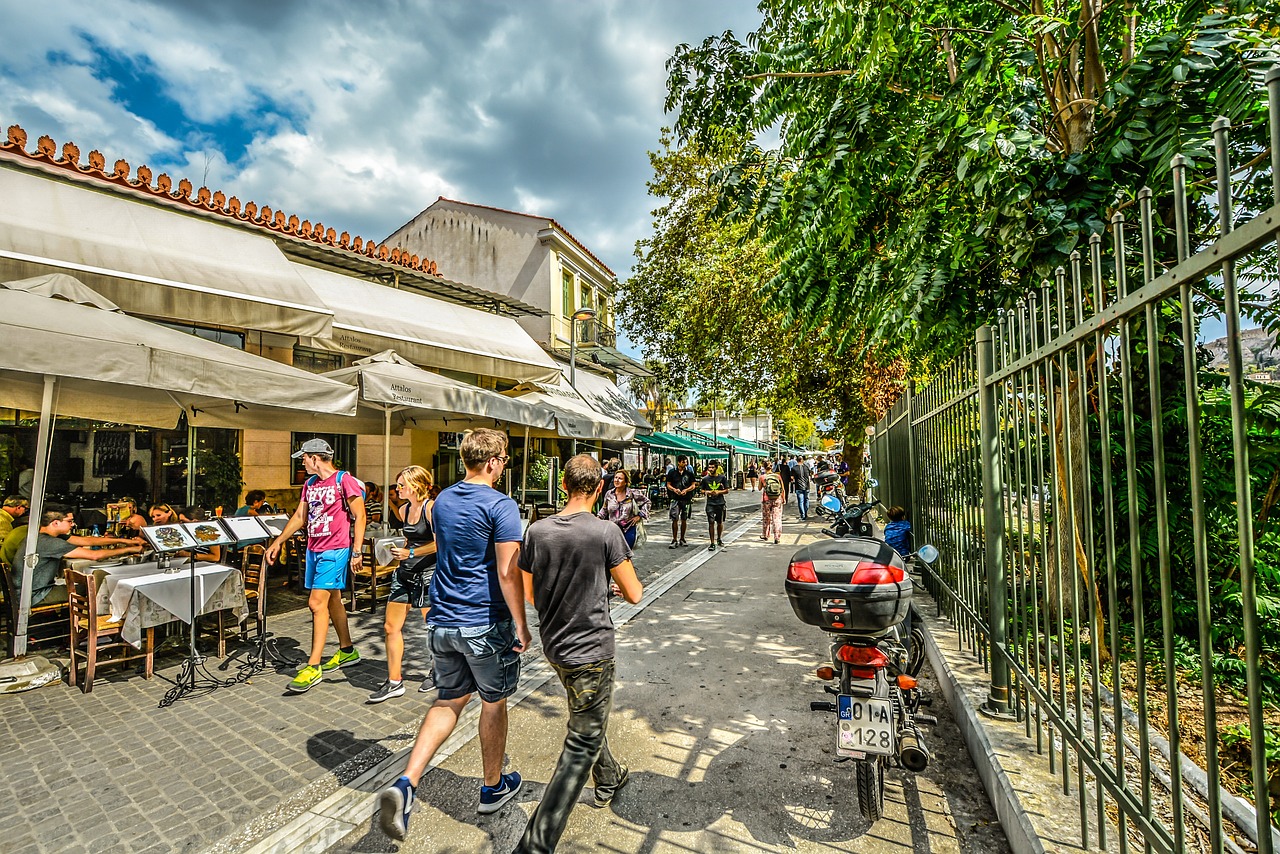 Pedestrians stroll along a sunny street lined with outdoor cafés and trees in Athens.