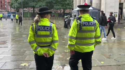 Two officers in police uniform including yellow reflective jackets, a woman with blonde hair and a man with short dark hair, walk across a paved square.