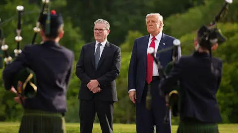 Getty Images Two young boy pipers in Highland dress frame either edge of the image, their backs to us. In the middle stands Sir Keir Starmer in dark blue suit, white shirt and patterned grey tie. His hands are crossed in front of him. To his left (our right) stands President Donald Trump. He wears a dark blue suit, white shirt and bright red tie. He has a stars and stripes badge pinned to his left lapel. They are both listening to the music and there are full green bushes behind them.