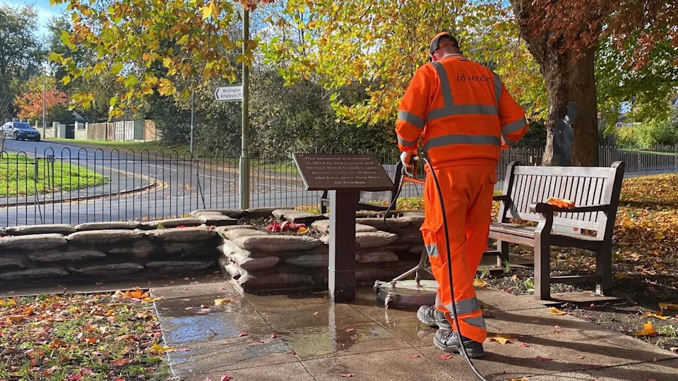 Mike pictured using a large, industrial pressure washer on flagstones next to the memorial, which is a cross-shaped flower bed with a bench next to it and a plaque. He's wearing a bright orange hi-vis jumper with silver reflective stripes, and a bright orange baseball cap. He's stood in a park, with the grass covered in orange leaves, and the leaves still on the trees are shades of red and orange. There's strong sunshine and the sky is blue.