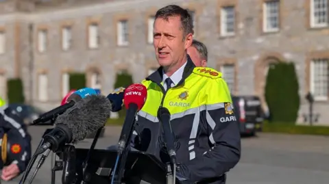 PA Media Garda Commissioner Justin Kelly addresses Tuesday's disorder in a statement to the media at Garda Headquarters in Dublin. He has short dark hair, wearing a white shirt and black tie and garda high viz jacket. He is standing in front of media microphones with a brick building behind him