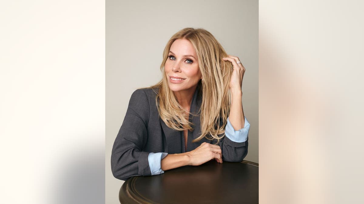 Cheryl Hines poses in a gray blazer with light blue cuffs, resting her arm on a table and smiling while touching her hair.