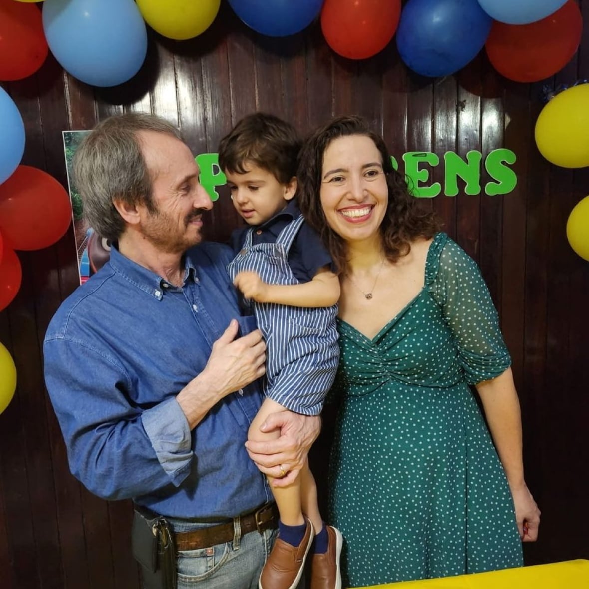 A family is pictured during what appears to be a birthday party, balloons mounted to the dark-coloured wall behind them. To the left, a man in denim shirt and jeans is carrying a young boy in striped blue-and-white overalls and a dark blue T-shirt. To the right, a woman in a dark green dress with small white polka dots.