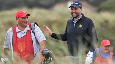 Getty Images JD Vance, sporting a white golf glove on his left hand, black fleece, blue shirt, grey trousers and blue Titleist cap, is talking to his caddy. The caddy is wearing a red Trump Turnberry cap and tabard, a spotted blue and white shirt, and has heavily tattooed arms. A second caddy, dressed in red hat and tabard and a blue shirt, is in the background. We can make out gorse and heather behind them all.