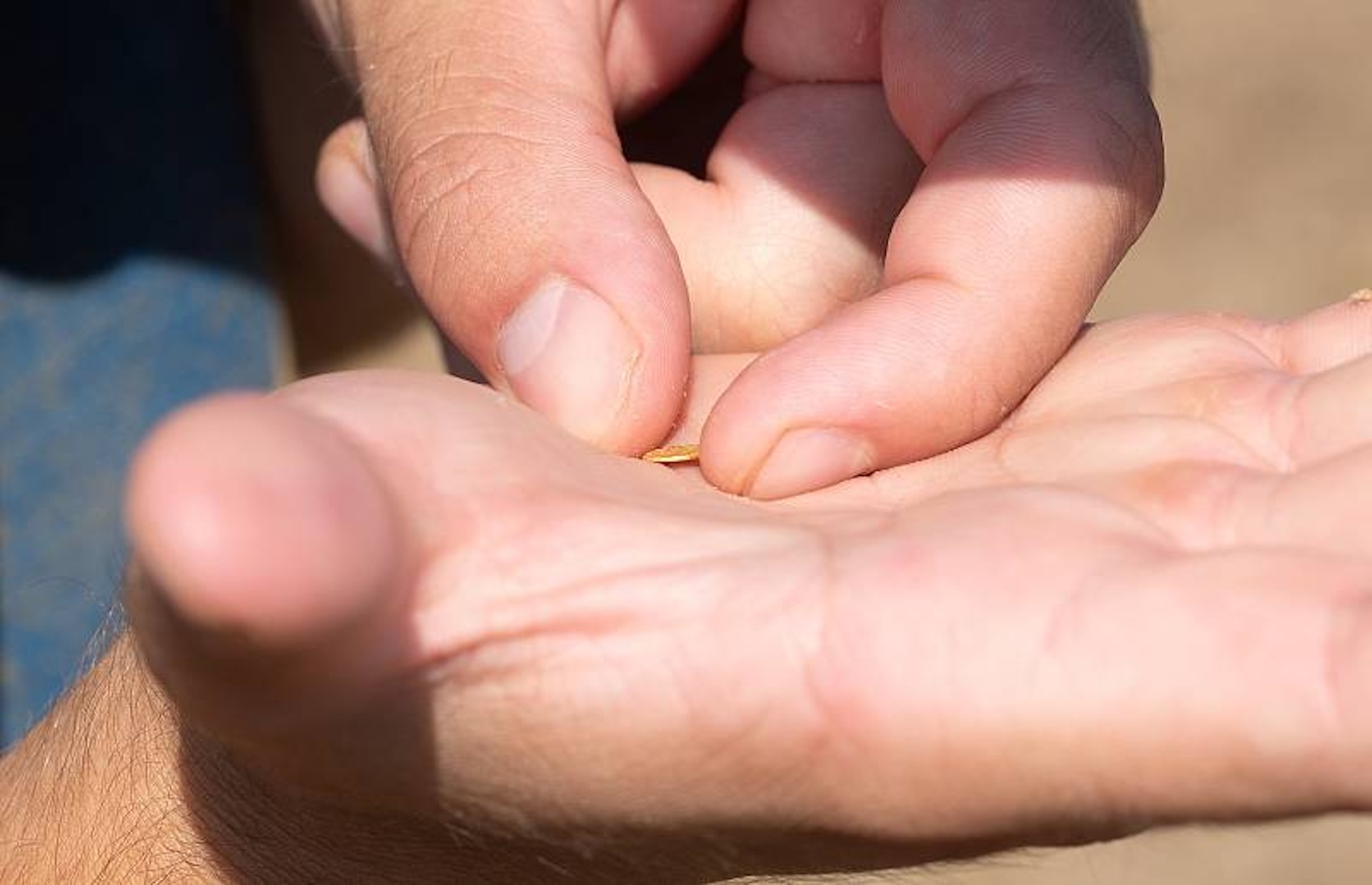 a close-up of a man's hands holding a tiny gold coin less than a centimeter wide