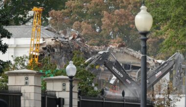 Demolition of East Wing of the White House on October 21, 2025. Construction machinery and the rubble and destroyed roofline of a white building can be seen over stone pillars and a black iron fence, with lampposts in the foreground.