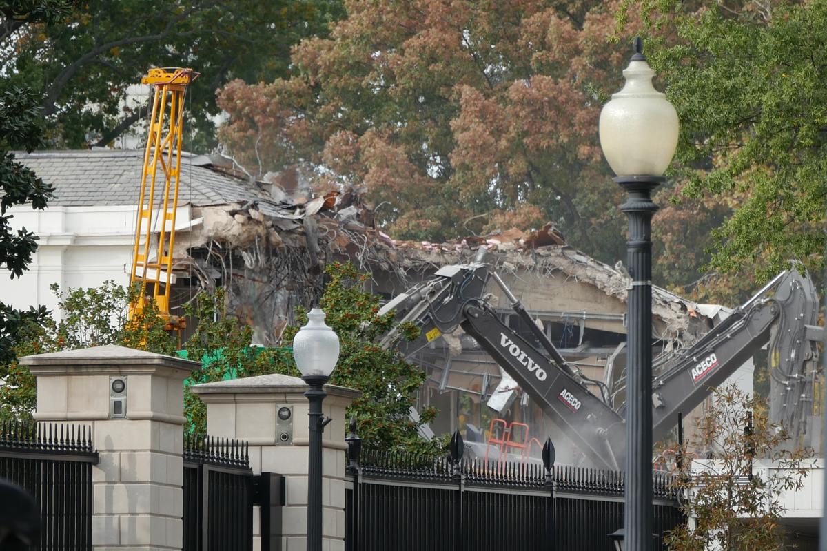 Demolition of East Wing of the White House on October 21, 2025. Construction machinery and the rubble and destroyed roofline of a white building can be seen over stone pillars and a black iron fence, with lampposts in the foreground.