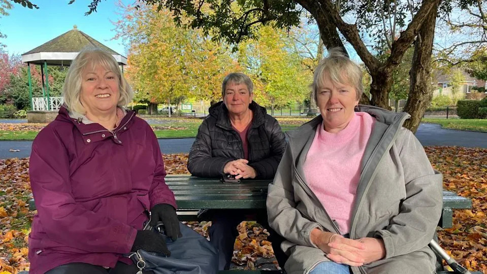 Three women sat together on a green park bench. Left to right; Georgina is wearing a maroon raincoat, Debbie is wearing a black raincoat, and Lynnette is wearing a pink jumper with a grey coat. The ground is covered in orange, fallen leaves, and you can see a green and white bandstand in the background. The leaves still on the trees are shades of red, green, and orange. There's strong sunshine and the sky is blue.