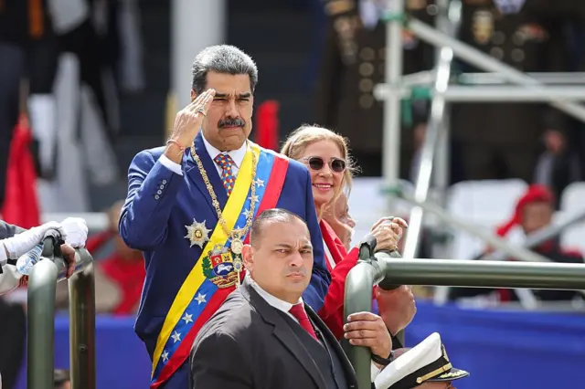 El presidente de Venezuela, Nicolás Maduro, vestido con un traje y una banda militar, saluda al llegar a un desfile militar como parte de las celebraciones del 214º aniversario de la independencia de Venezuela el 5 de julio de 2025 en Caracas, Venezuela. Un hombre se encuentra frente a él sosteniendo una barra y una mujer sonríe detrás de él.