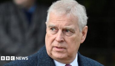 Prince Andrew has short grey hair and is gazing to one side. He is wearing a dark blue jacket, a white shirt and a red tie.