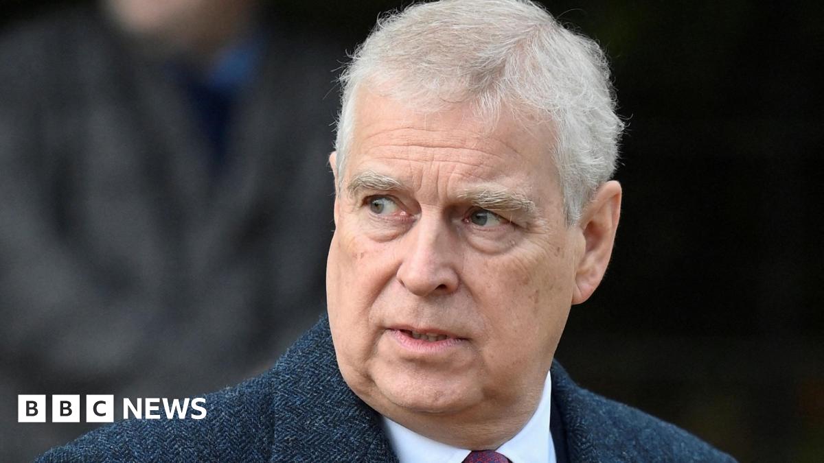 Prince Andrew has short grey hair and is gazing to one side. He is wearing a dark blue jacket, a white shirt and a red tie.