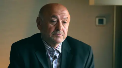 A man wearing a white shirt with red and blue stripes with a smart dark jacket looks at the camera. He has a greying moustache, short facial hair and is bald.