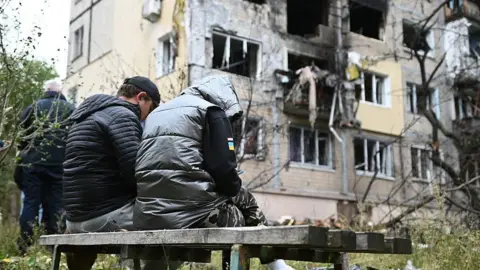 Getty Images Two people sit on a bench against the backdrop of a destroyed building