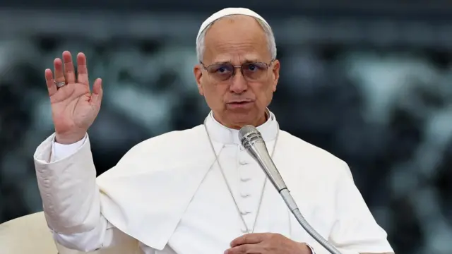 Pope Leo in a white tunic with his hand up, speaking into a microphone.