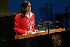 A dark-haired woman in a red jacket speaks into a microphone.