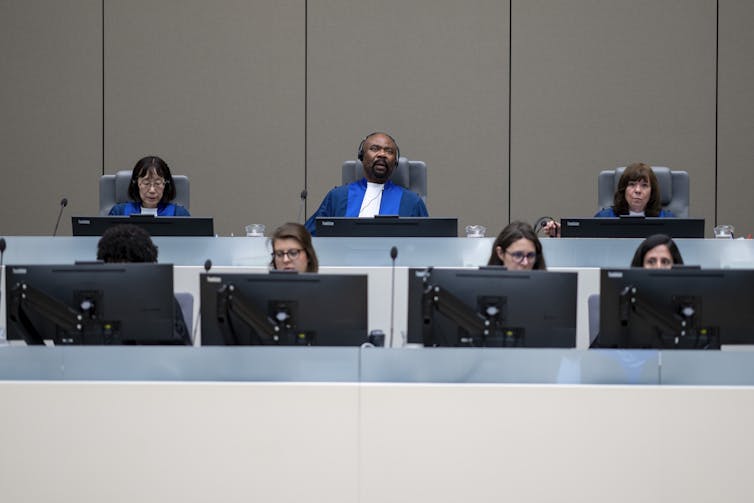 Three judges in blue robes sit in a row behind people looking at computer screens.