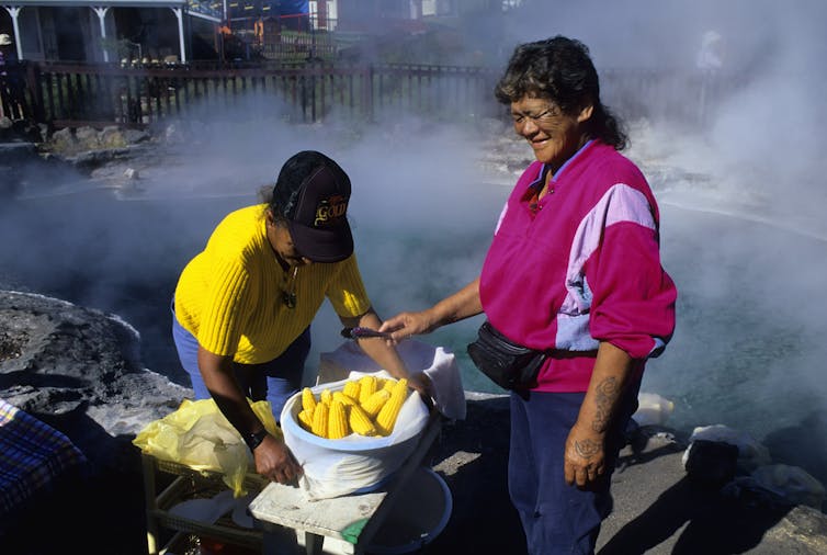 Two people stand near a large container of shucked corn while steam billows from a pool of water behind them.