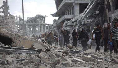 Mehrere Menschen laufen durch eine Straße voller Schutt und eingestürzter Gebäude. Im Hintergrund sind schwer beschädigte Betonbauten zu sehen.