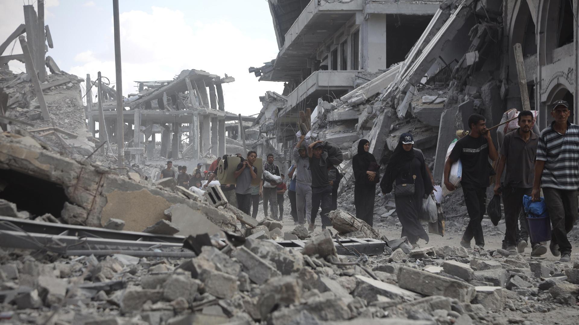 Mehrere Menschen laufen durch eine Straße voller Schutt und eingestürzter Gebäude. Im Hintergrund sind schwer beschädigte Betonbauten zu sehen. Mehrere Menschen laufen durch eine Straße voller Schutt und eingestürzter Gebäude. Im Hintergrund sind schwer beschädigte Betonbauten zu sehen.