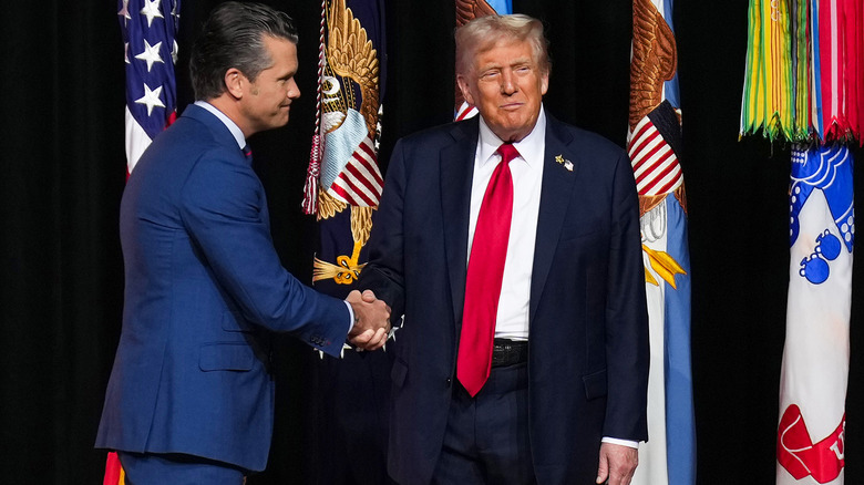 Pete Hegseth shaking Donald Trump's hand in front of some flags while both wear suits