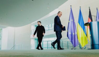 Friedrich Merz and Volodymyr Zelenskyy walk past EU, Ukrainian, and German flags after a press conference.