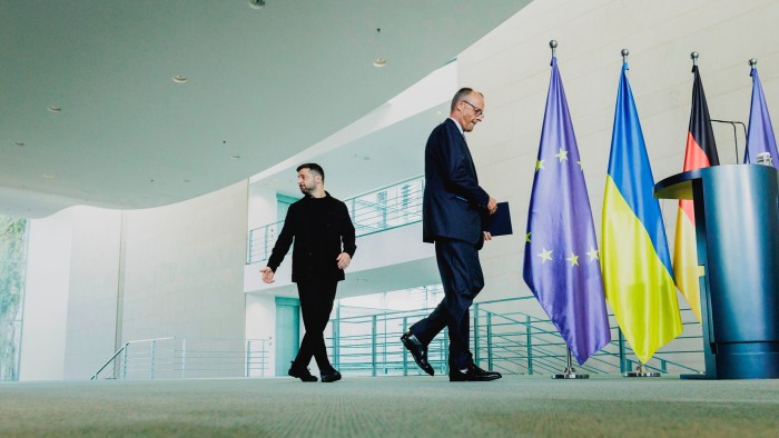 Friedrich Merz and Volodymyr Zelenskyy walk past EU, Ukrainian, and German flags after a press conference.