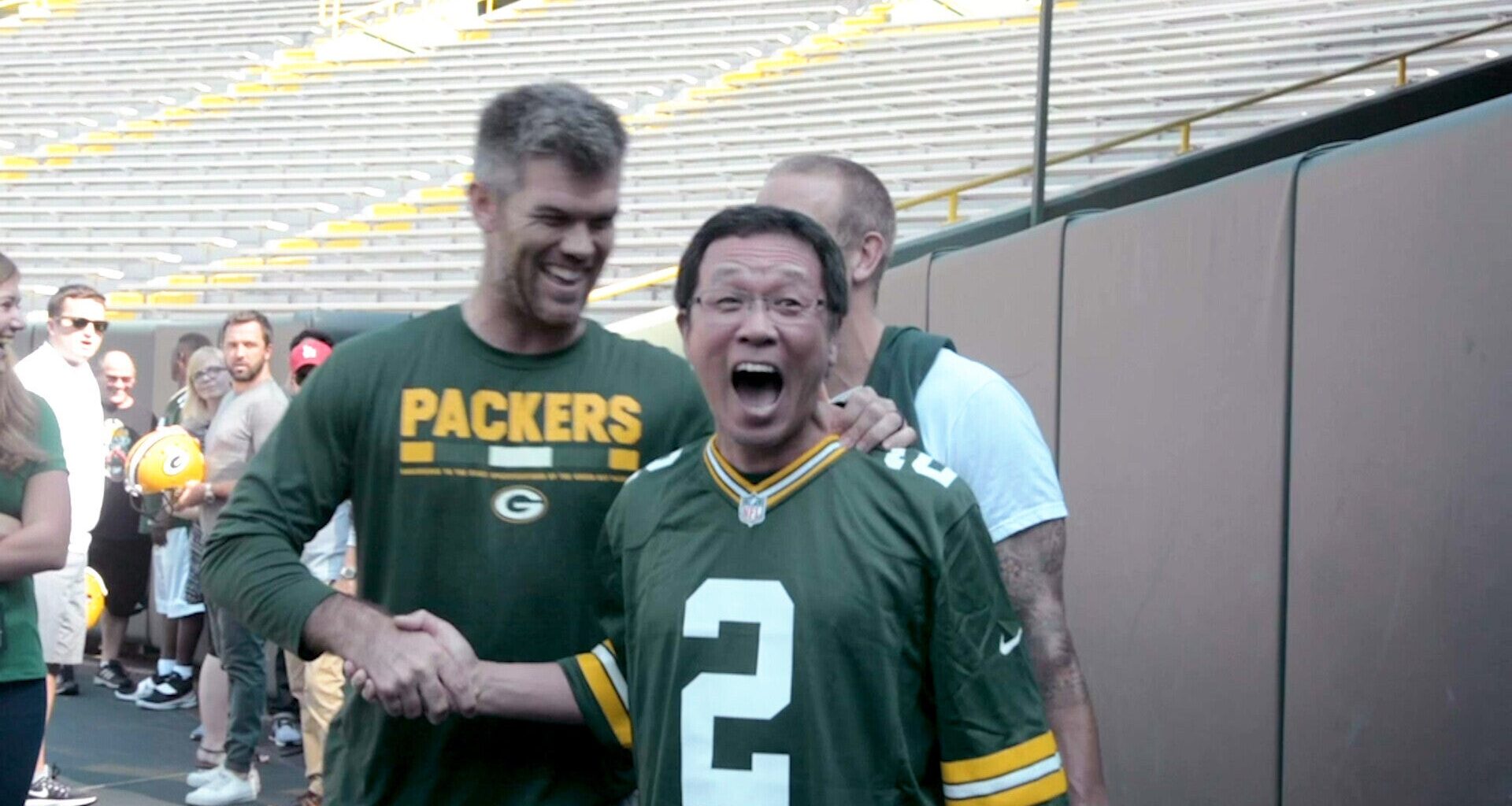 Two men in Green Bay Packers jerseys smile and shake hands on a football field, with empty stadium seats in the background.