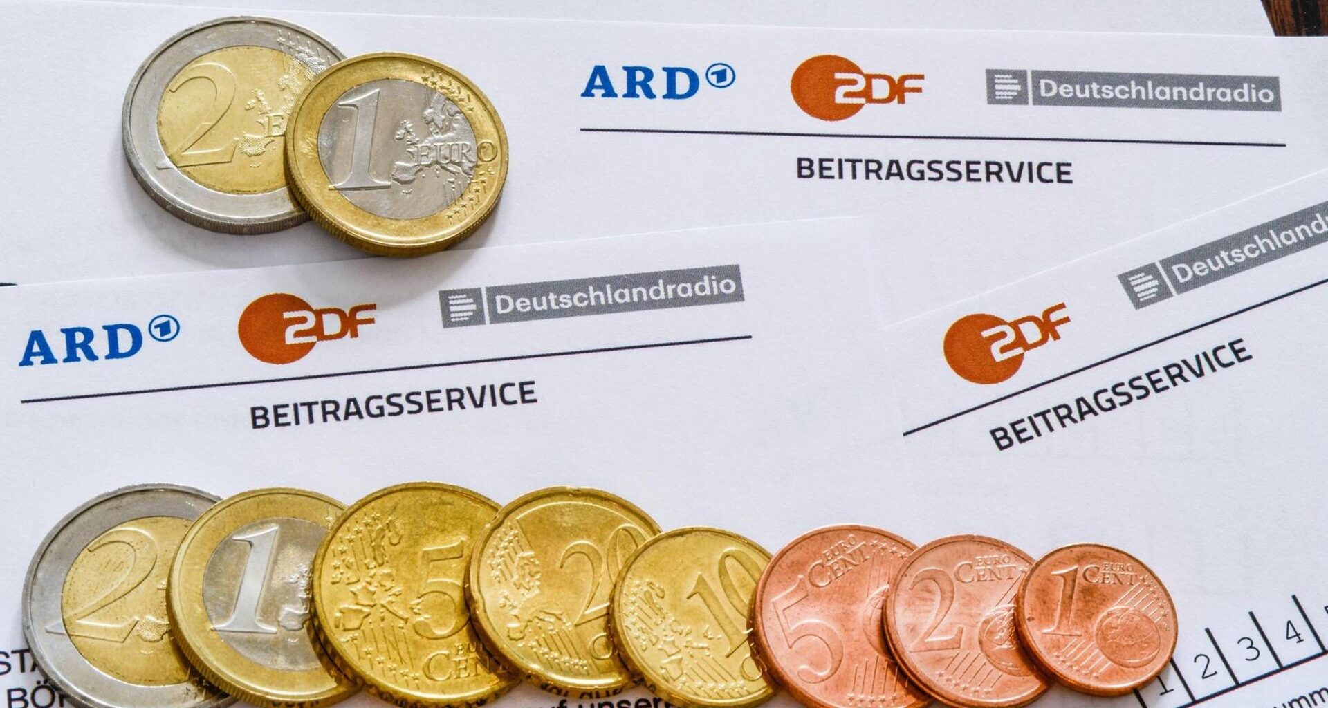 Auf mehreren Formularen des Beitragsservice für den Rundfunkbeitrag liegen Geldmünzen.